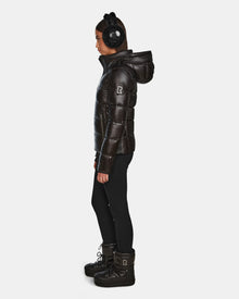 A woman in profile wears black earmuffs, black leggings, and winter boots, showcasing the Rudsak MIRRY S - L123545 Black quilted down coat with hood, set against a plain white background.