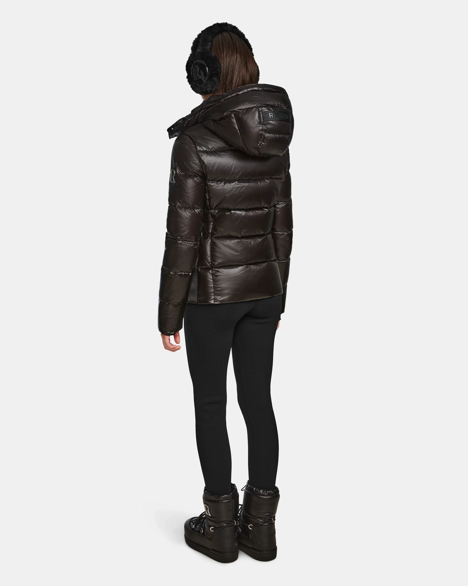 A person stands facing away wearing black earmuffs, black leggings, black winter boots, and a shiny Rudsak MIRRY S - L123545 Black womens quilted down coat with hood against a plain white background.