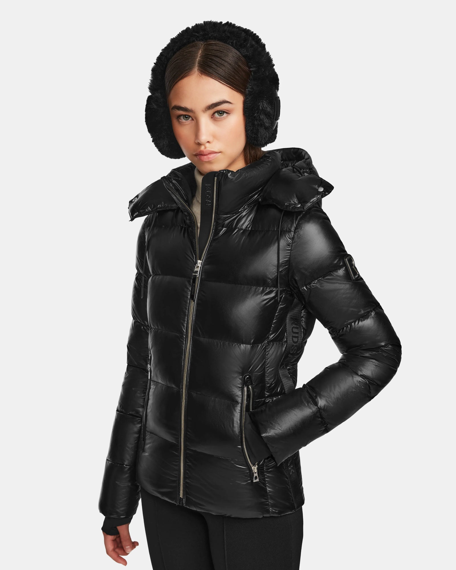 A woman in a shiny black Rudsak MIRRY S - L123545 winter coat with a hood and black earmuffs stands against a plain light background, looking slightly to the side with a neutral expression.