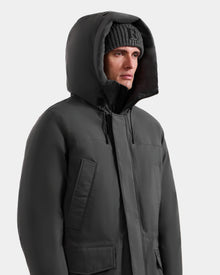 A man in the MITCHELL - M125513 Grey men’s down parka by Rudsak, filled with RDS-certified down, and a ribbed knit beanie, gazes to the side against a plain light background.