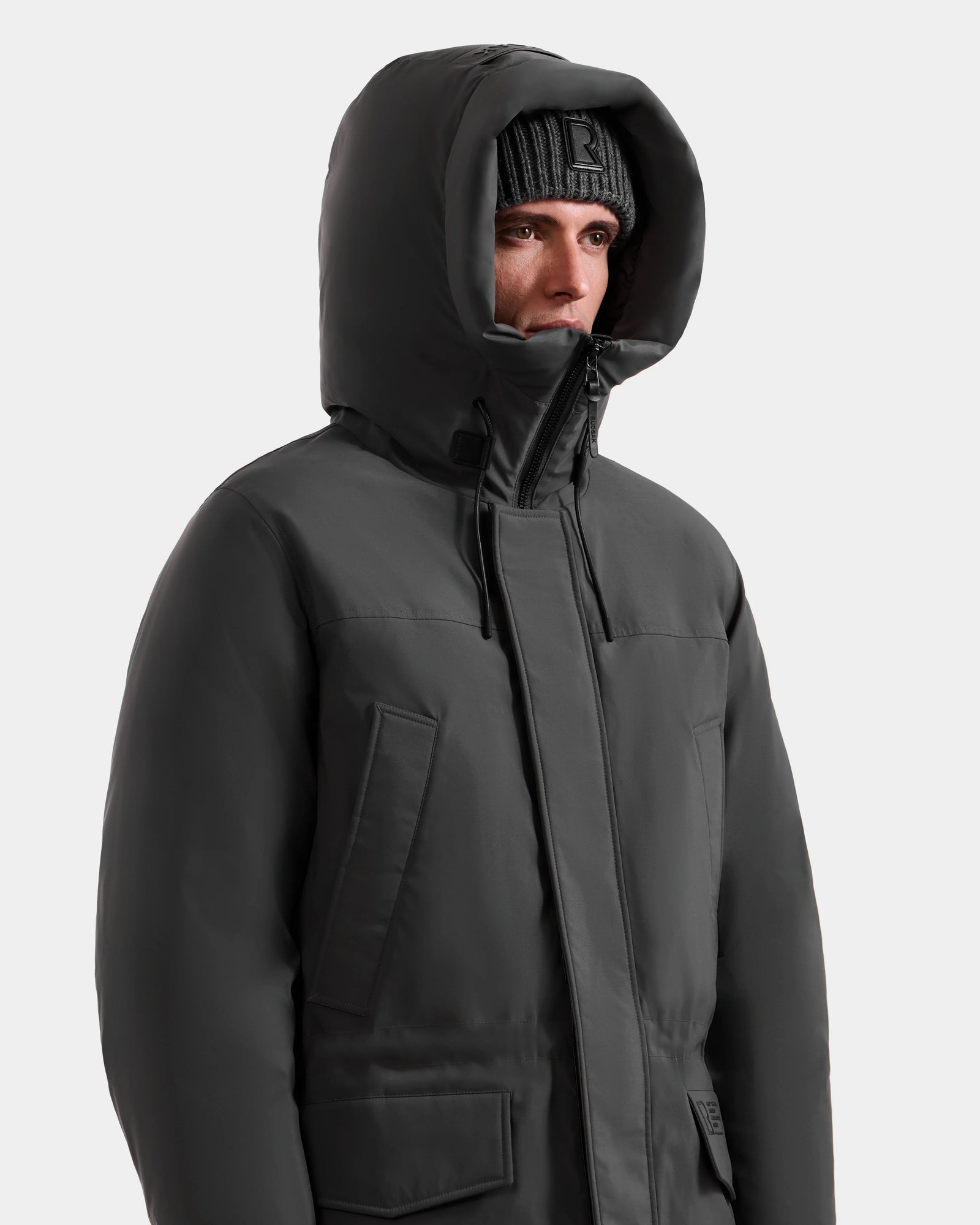 A man wearing the Rudsak MITCHELL - M125513 Grey down parka and a knit beanie looks off to the side against a plain, light background.