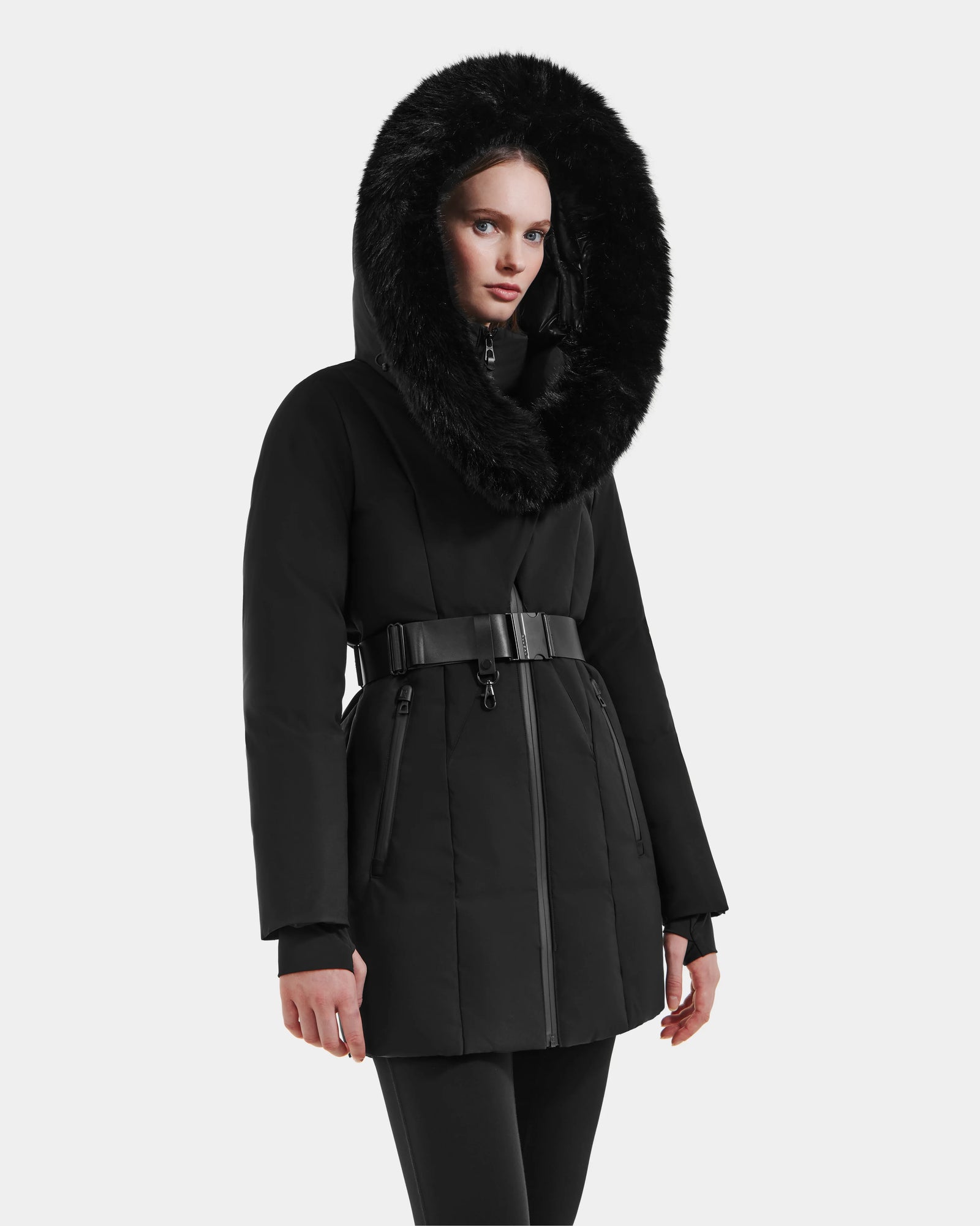 A woman wears the Rudsak MODA M - L125555 Black down puffer coat with a large fur-lined hood and vegan leather belt, standing against a plain white background.
