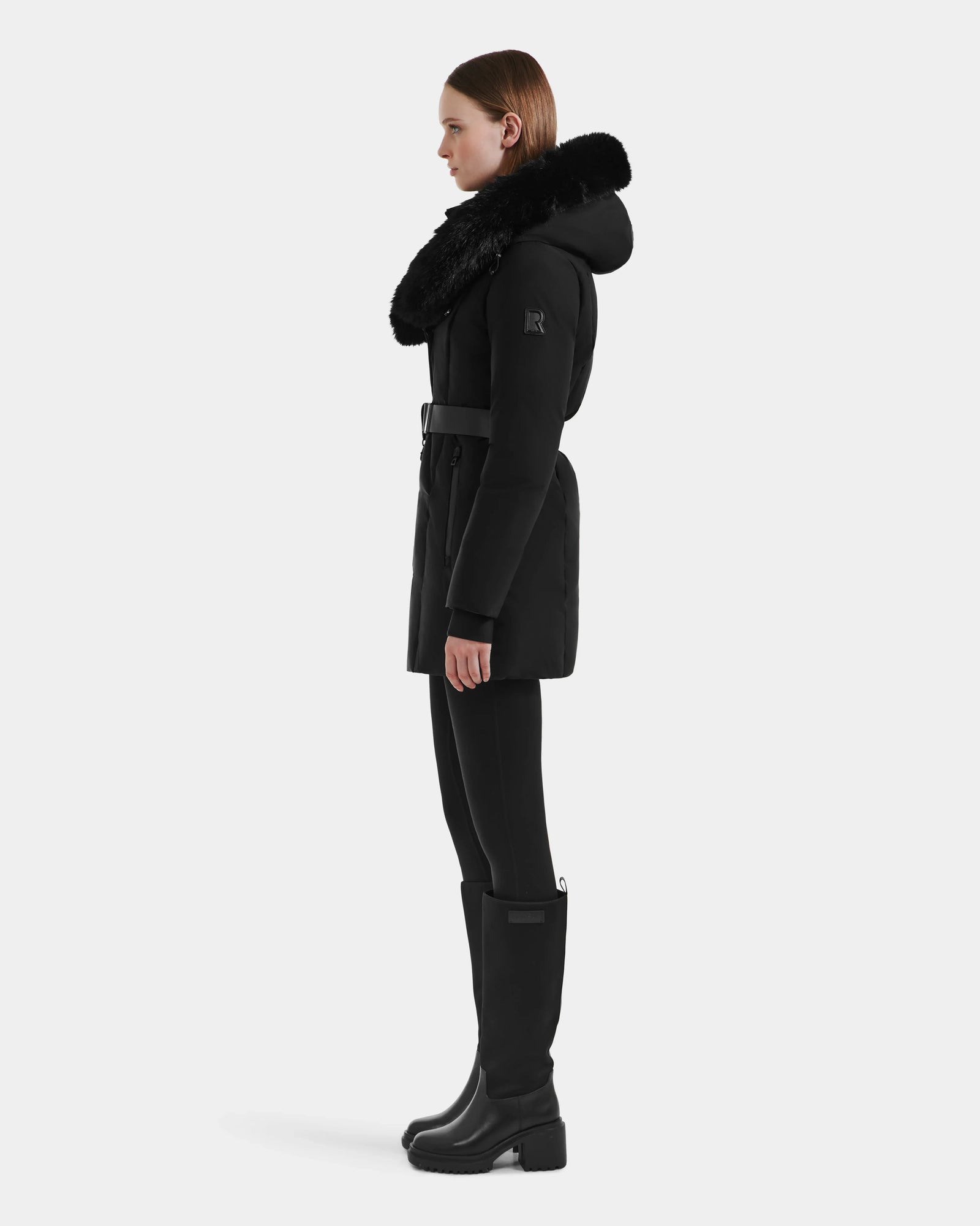 A woman stands in profile against a light background, wearing the Rudsak MODA M - L125555 Black winter coat with a large faux fur-trimmed hood, black leggings, knee-high boots, and a vegan leather belt at the waist.
