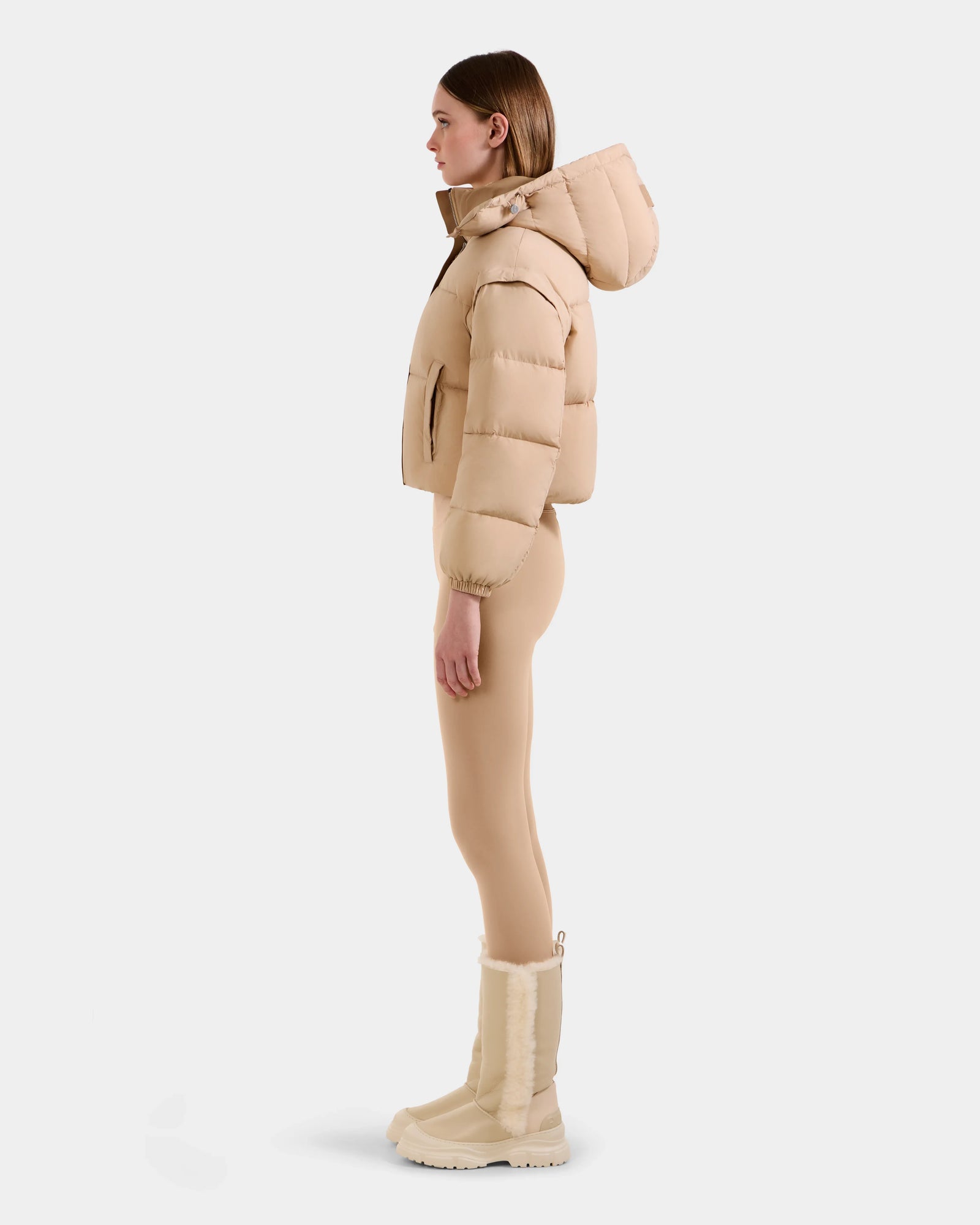 A woman stands in profile wearing the Rudsak OPRAH - L125546 Vanilla cropped down puffer with removable sleeves, paired with beige fitted leggings and beige fur-trimmed winter boots, set against a plain light background.