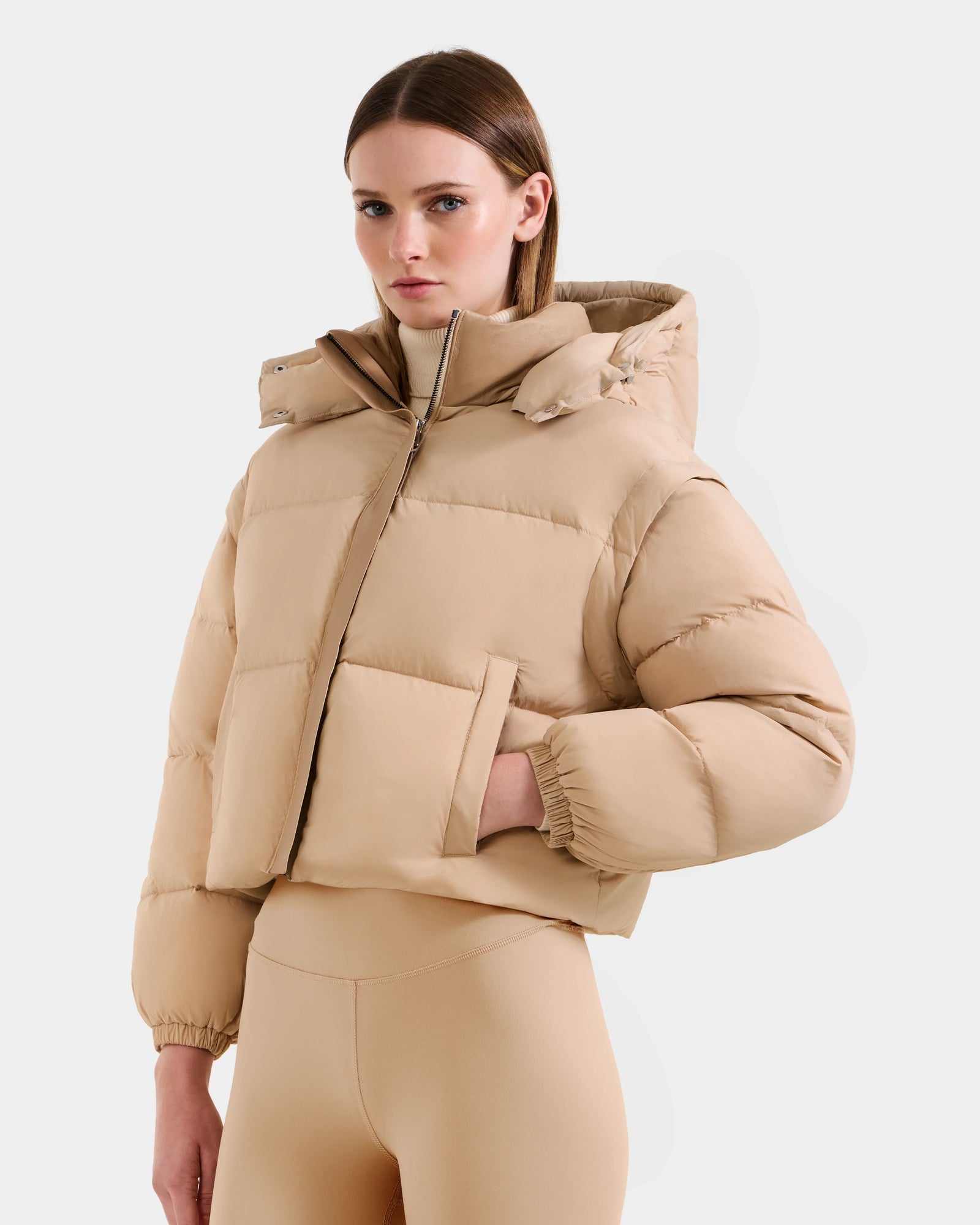 A woman stands against a plain background wearing the Rudsak OPRAH - L125546 Vanilla jacket, featuring a high collar and hood, paired with matching beige leggings. She looks confidently at the camera with one hand in her pocket.