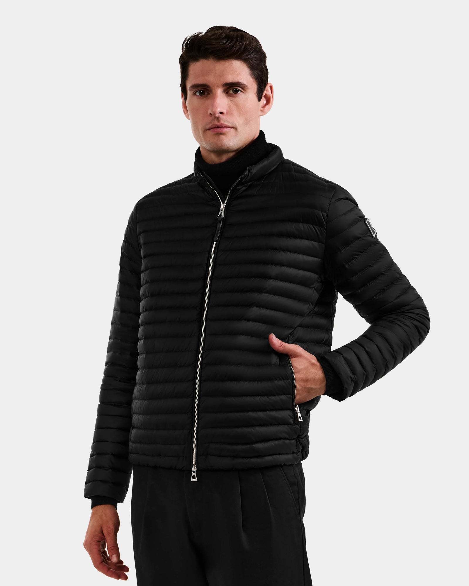 A man stands against a plain white background, his left hand in the pocket of the Rudsak REYMOND M - M125523 Black men's down jacket, made with RDS-certified down and recycled nylon.