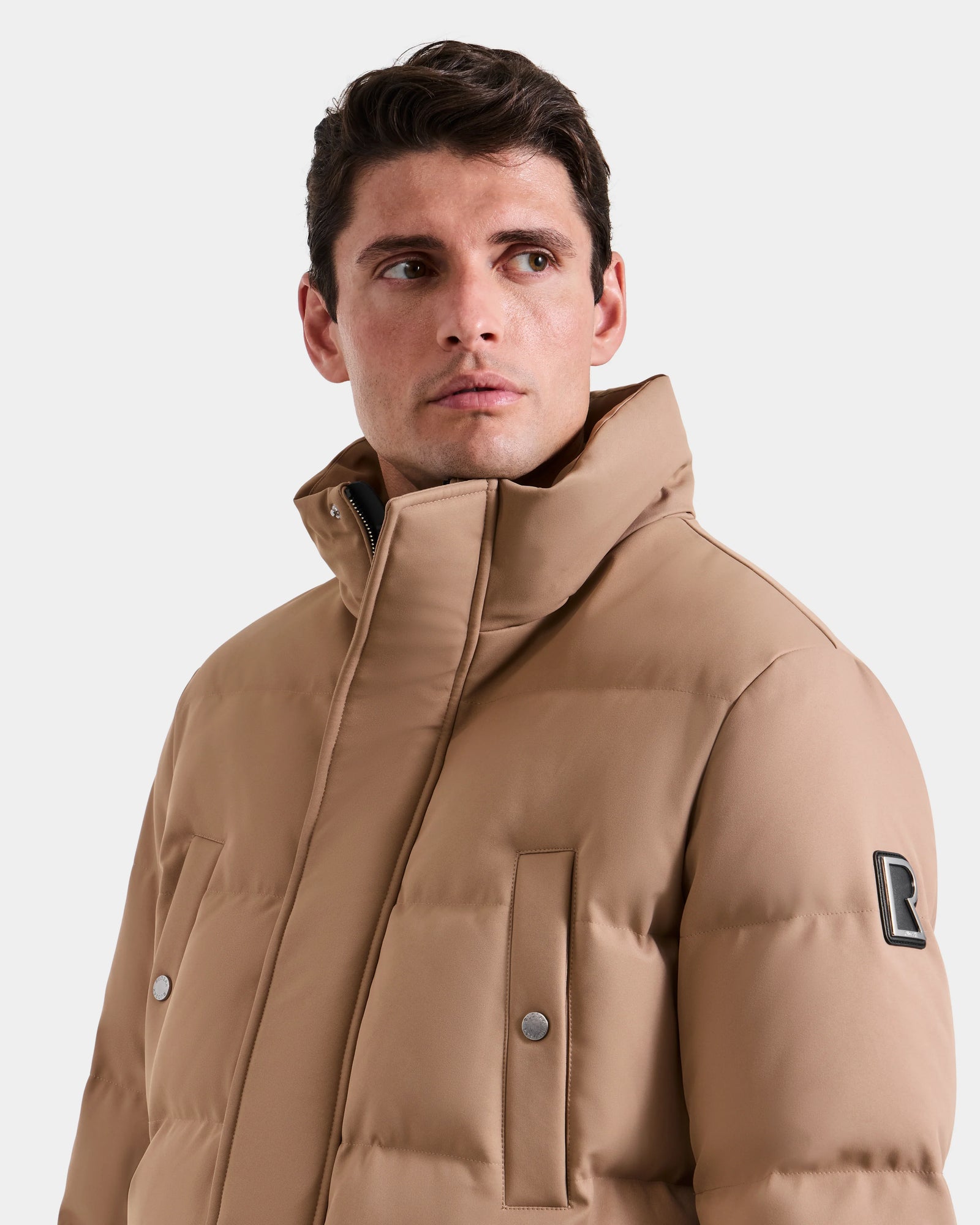 A man wearing the Rudsak RICKY - M123502 Sand men’s quilted down parka with a high collar looks to the side. The jacket features snap-button pockets and a sleeve patch, standing out against the plain white background.