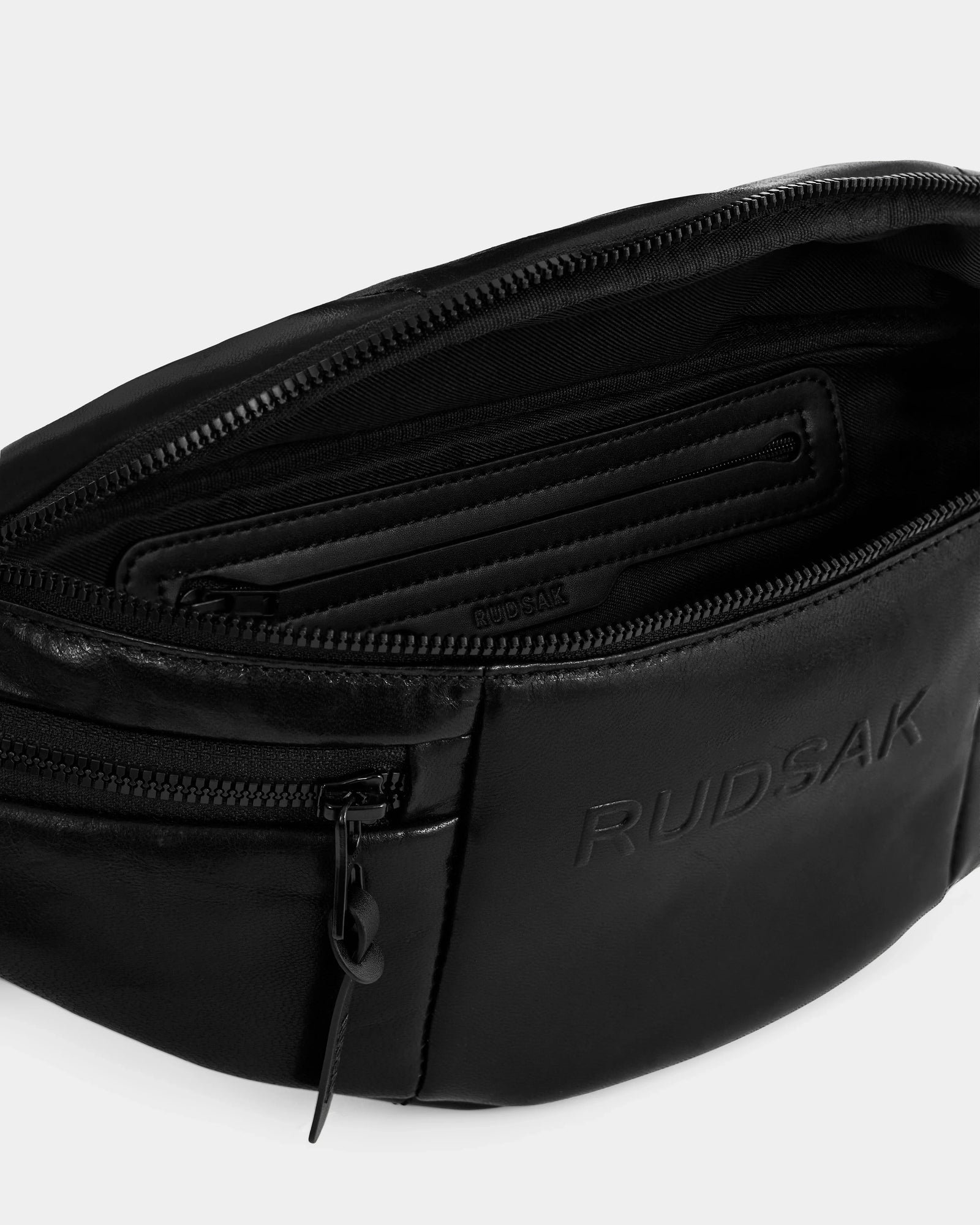 Close-up of RILONI L - U325002 Black by Rudsak, an open black leather belt bag featuring a zippered main compartment, inner zip pocket, and the embossed brand name—a stylish unisex fanny pack for daily essentials.