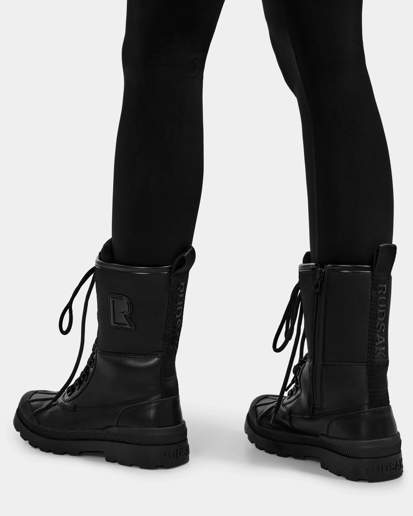 A person is seen from the back against a white background, wearing black leggings and Rudsak RITA - L222502 Black lace-up combat boots made with weather-treated leather.
