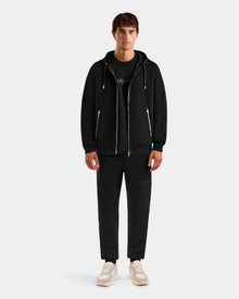 A young man faces forward, wearing the Rudsak ROGER - U523507 Black M unisex zip-up hoodie with black pants, a black T-shirt, and white sneakers, showcasing effortless urban style against a plain light background.