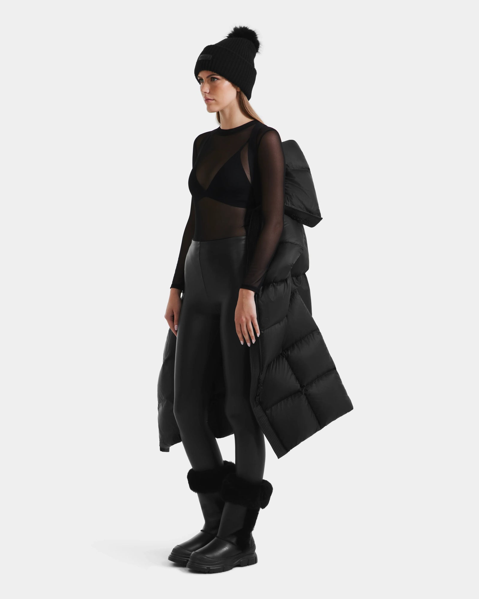 A woman stands against a plain background wearing the Rudsak SASHA - 8125686 Black womens long down puffer coat with removable hood draped over her shoulder, paired with a black beanie, sheer top, bra, leggings, and boots.