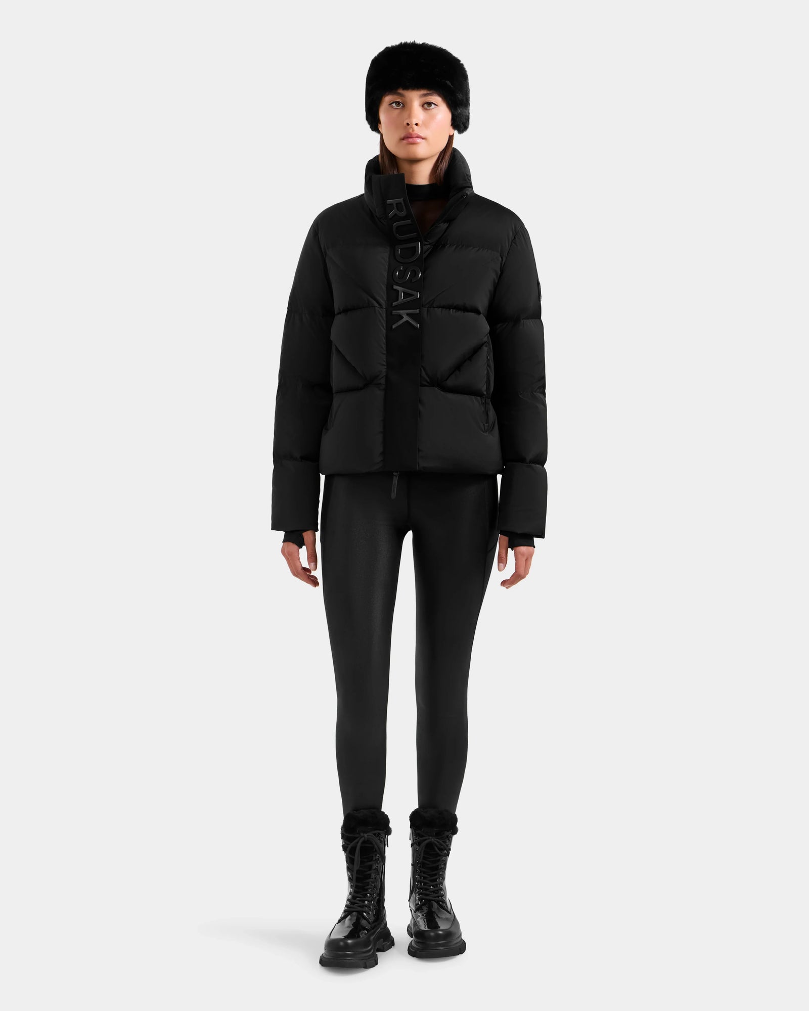 A person in the Rudsak STEFANIE M - L125526 Black puffer jacket, black leggings, lace-up boots, and a black fur hat stands facing forward against a plain light background.
