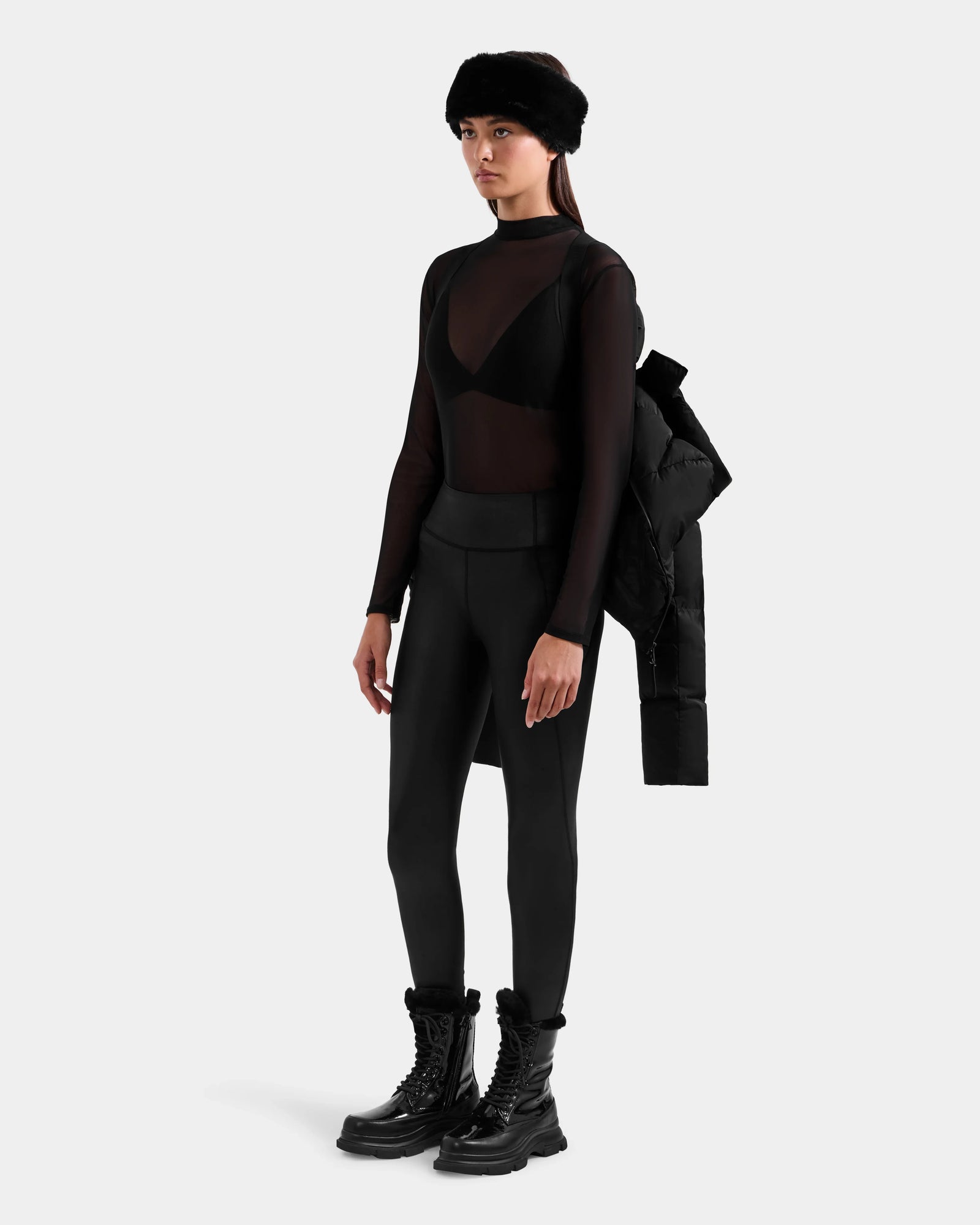 A woman models the Rudsak STEFANIE M - L125526 Black puffer jacket, draped over her shoulder, paired with black boots, fitted pants, a sheer long-sleeve top over a black bra, and a black fur headband against a plain light gray background.