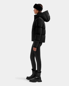 A person stands in profile against a plain light background, wearing the Rudsak STEFANIE M - L125526 Black puffer jacket with a hood, paired with black leggings, chunky black boots, and a black headband.