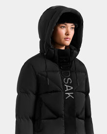 A woman with straight dark hair and a neutral expression stands against a plain light background, wearing the Rudsak STEFANIE M - L125526 Black quilted down jacket with a removable hood and vertical RUDSAK lettering.
