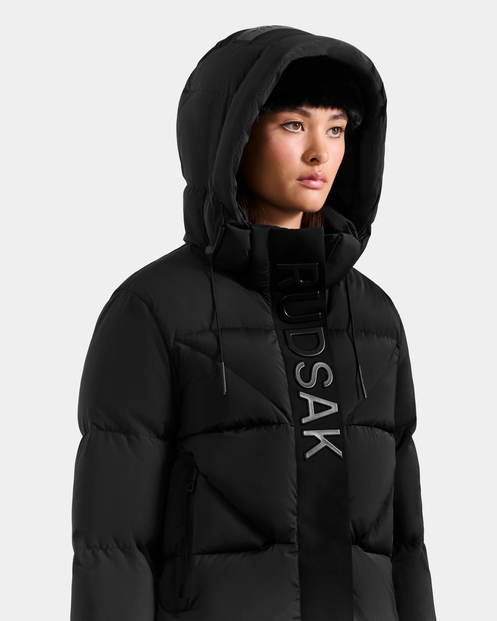 A woman with straight dark hair and a neutral expression stands against a plain light background, wearing the Rudsak STEFANIE M - L125526 Black quilted down jacket with a removable hood and vertical RUDSAK lettering.