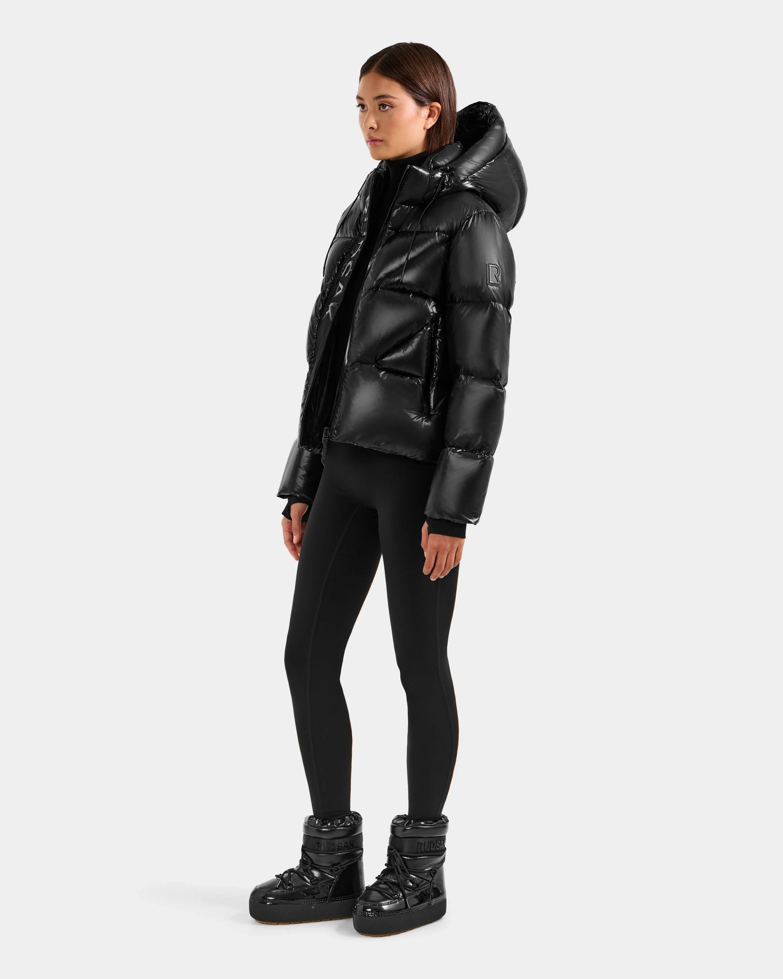A person stands against a plain light gray background wearing the Rudsak STEFANIE S - L125585 Black quilted down puffer with a hood, paired with black leggings and black padded ankle boots.