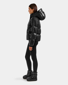 A woman stands in profile wearing the Rudsak STEFANIE S - L125585 Black, a shiny women's short puffer jacket with a hood, paired with black leggings and winter boots against a plain light gray background.