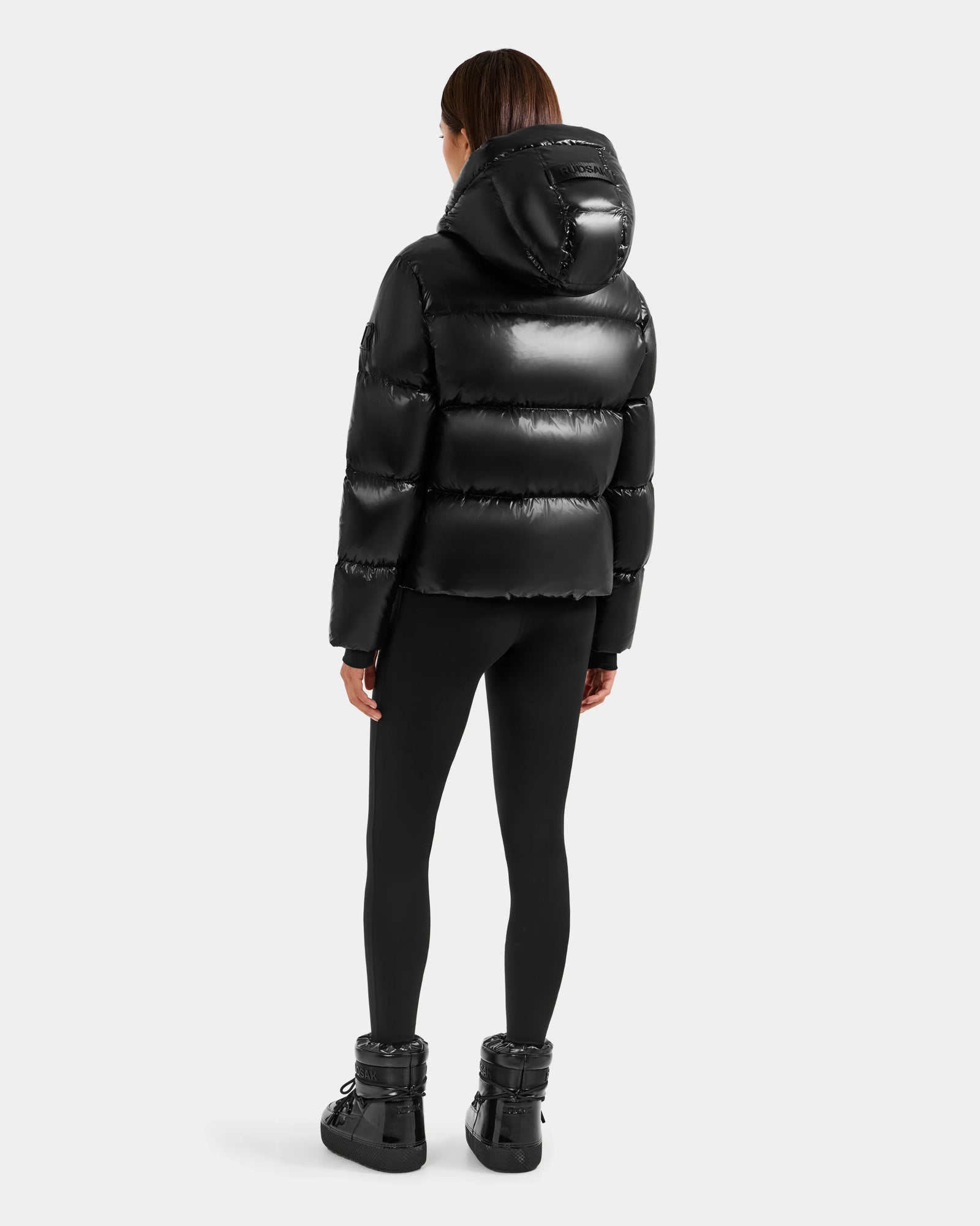 A person with straight brown hair, seen from behind, wears the Rudsak STEFANIE S - L125585 Black shiny puffer jacket with hood, black leggings, and black winter boots against a plain light gray background.