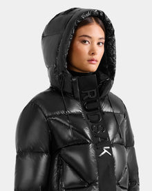Wearing the STEFANIE S - L125585 Black weather-resistant quilted down puffer jacket by Rudsak, with its hood up and a bold black RUDSAK scarf, a person poses against a light grey background.