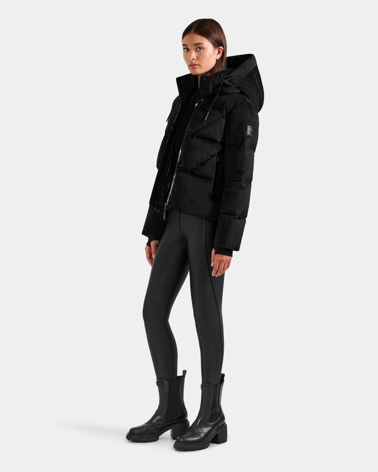 A woman wears the Rudsak STEFANIE V - L125584 Black down puffer jacket with hood, paired with black leggings and ankle boots, standing against a plain light background. She faces forward, hands by her sides, with a neutral expression.