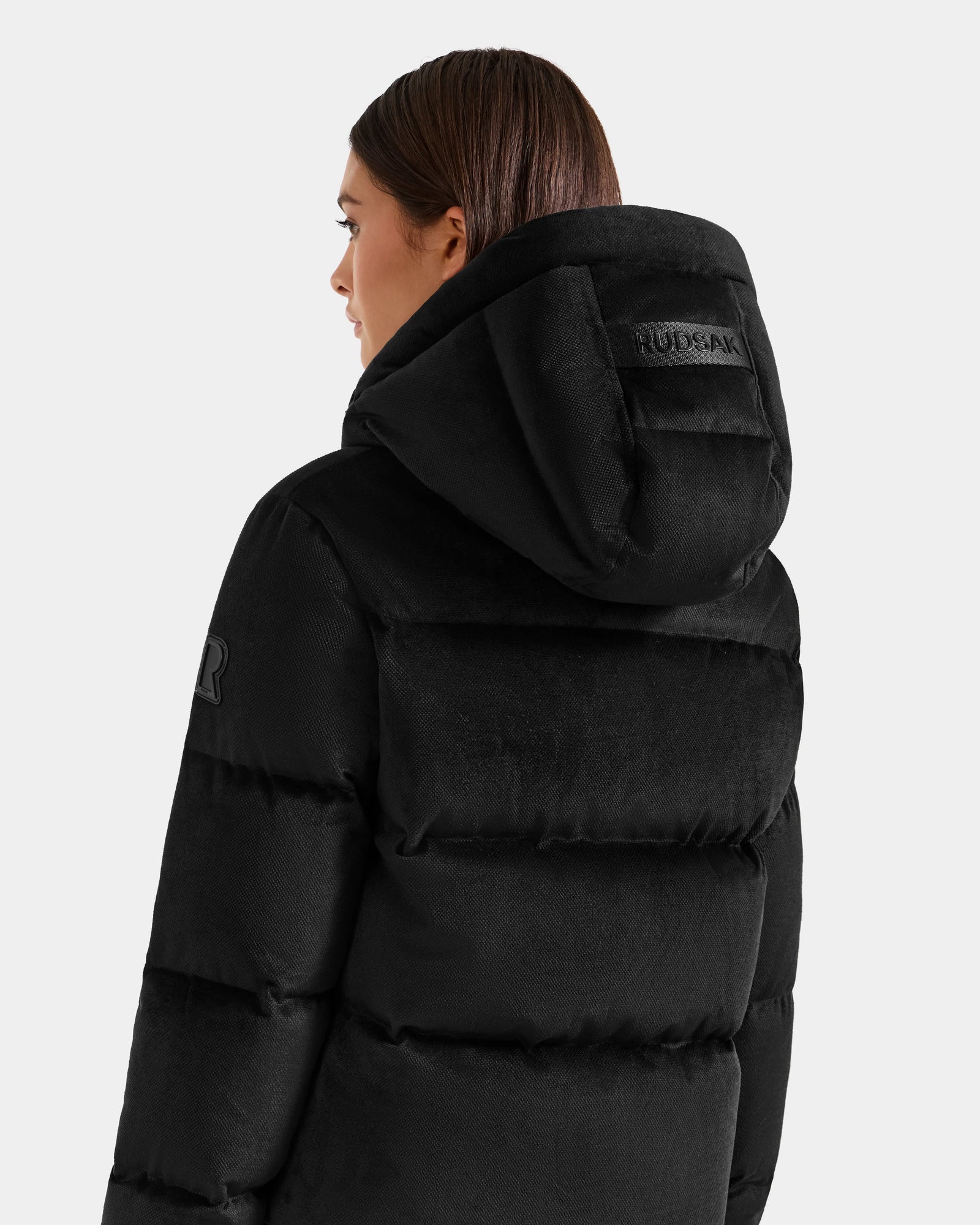 A person with straight brown hair is seen from behind wearing the Rudsak STEFANIE V - L125584 Black down puffer jacket with a hood and sleeve patch. The RUDSAK label is on the hood. Background: plain light gray.