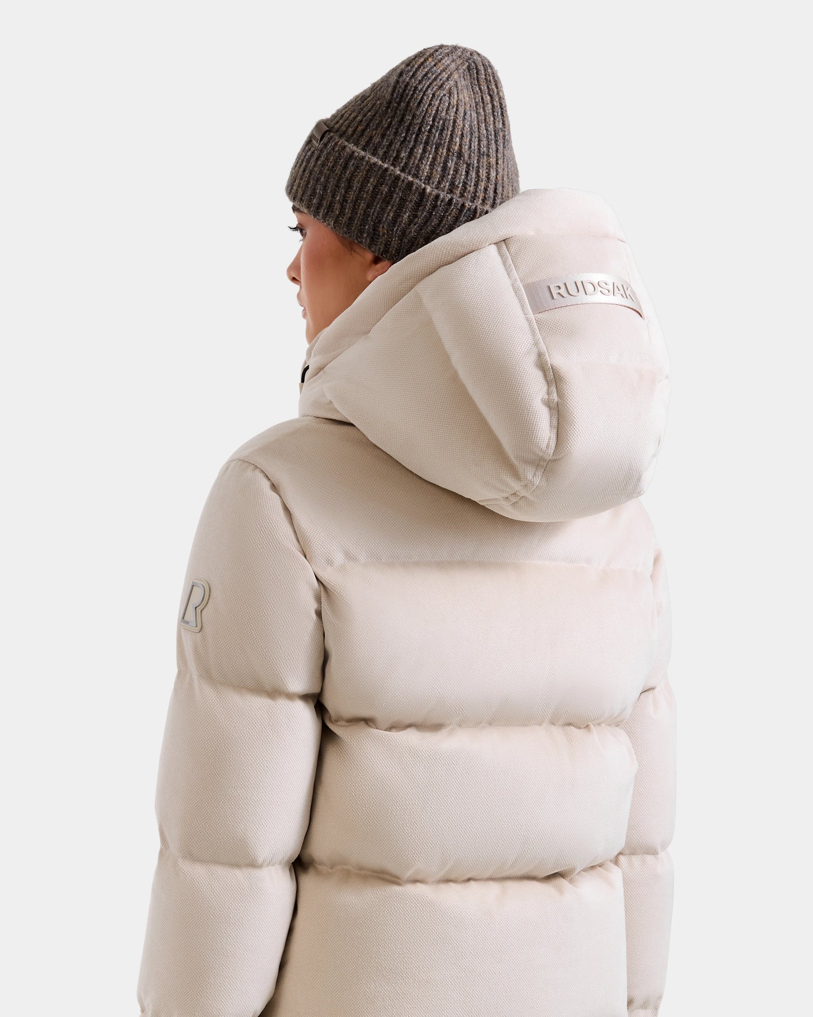 A person is shown from the back wearing the Rudsak STEFANIE V - L125584 Vanilla down puffer jacket with a hood and a brown knit beanie. The winter coat features Rudsak branding on the hood and an R patch on the sleeve.