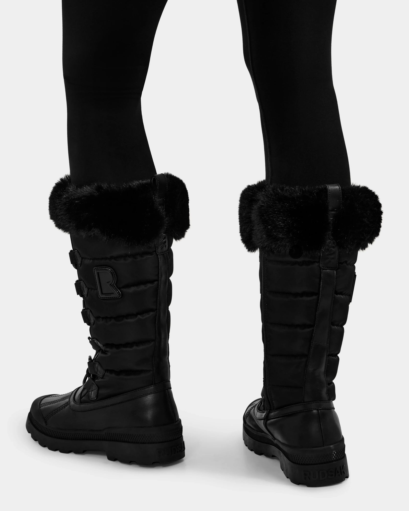 Someone stands on a white background wearing Rudsak's WINDY - L224507 Black women's knee-high winter boots, featuring faux fur trim, lace-up backs, quilted texture, and branding on the heel and side for cold-weather comfort.