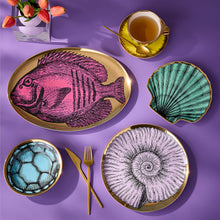 A styled flat-lay of a dinnerware set on a purple background, featuring a large oval platter with a pink fish illustration, a scallop-shaped dish in teal, a bowl with a blue soccer pattern, and two round plates with gold-leaf-edged nautilus and spiral designs. Gold cutlery, a yellow teacup, and a vase of tulips are arranged around them.