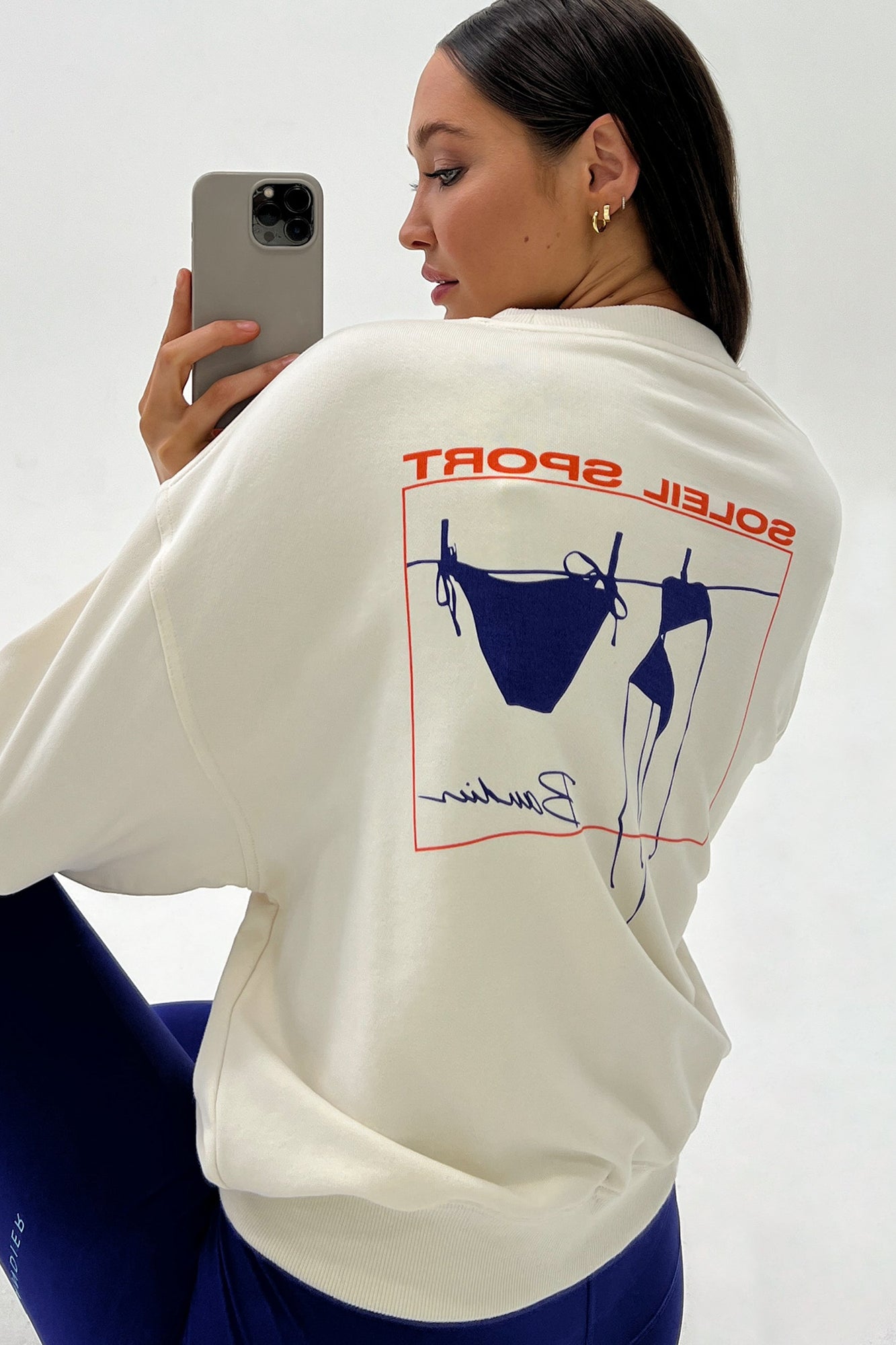 Model wears a white crewneck sweatshirt with logo on back that reads "Soleil Sport" and underneath is a square orange border containing a blue bikini hanging to dry on a clothesline. 