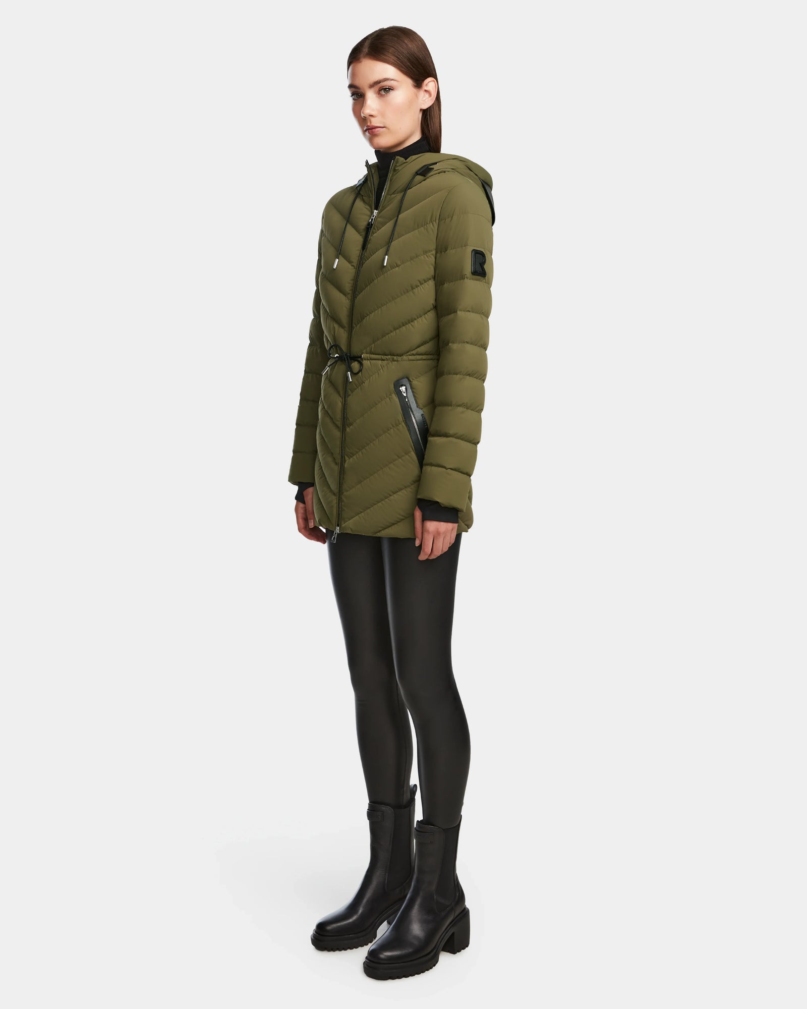 A woman wears the Rudsak FRANCINE - 8124722 Loden Green puffer jacket with black zipper details, paired with black leggings and boots, posed against a plain white background.