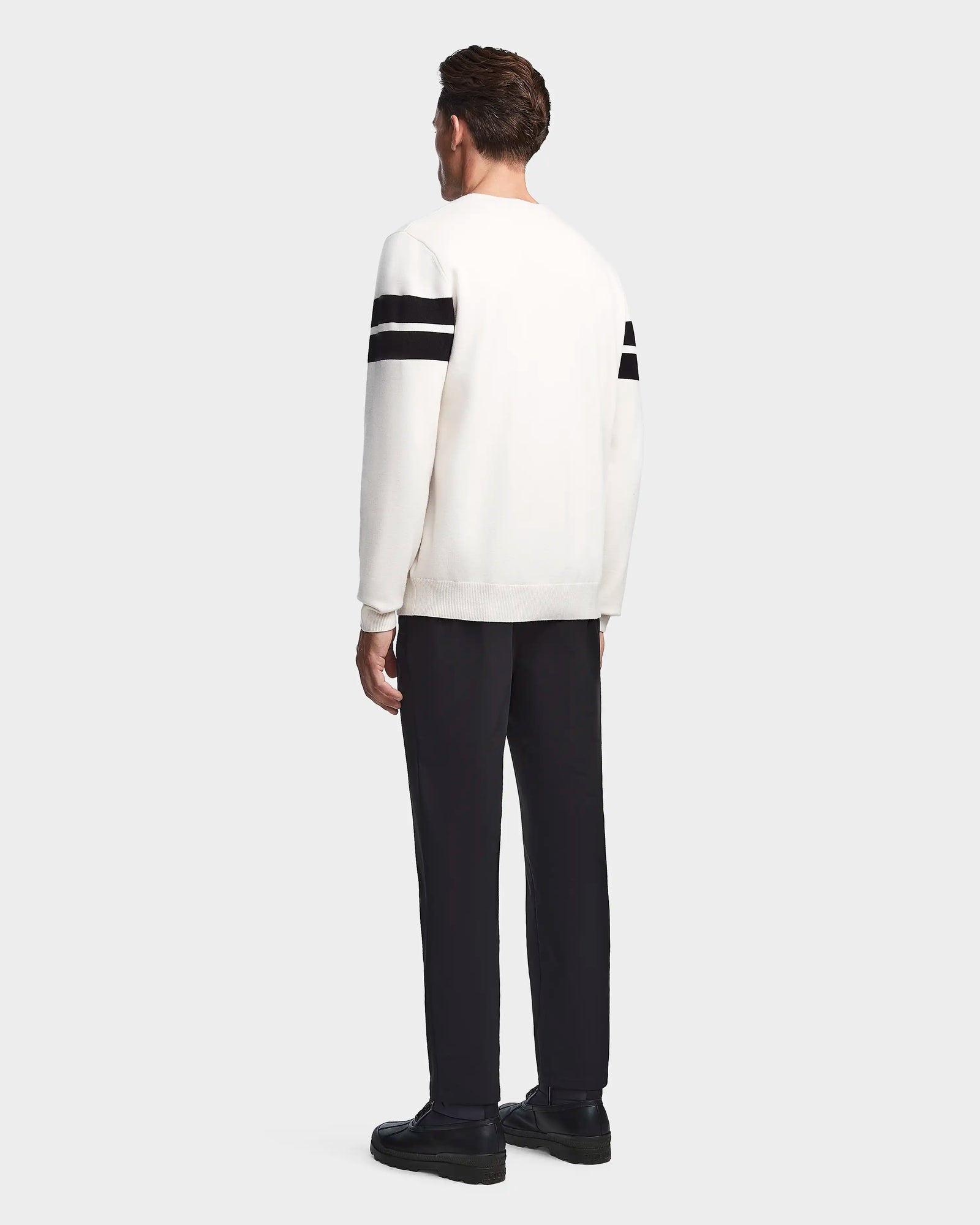 A man faces away, wearing the Rudsak OMAR - 6523507 Iceland white men’s knit sweater with two black stripes on each sleeve, paired with black pants and shoes, against a plain light gray background.