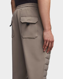 A person wearing Rudsak’s MIKE - 6523503 Cement men’s quilted pants, featuring taupe color, large flap back pockets, black pull tab, and padded side detailing. Only the lower back and one hand are visible.