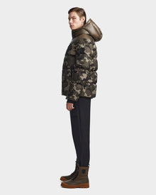 A person stands in profile wearing the Rudsak RONNY M - M123512 Camouflage weather-resistant jacket with a hood, paired with black pants and brown lace-up boots, set against a plain light background.