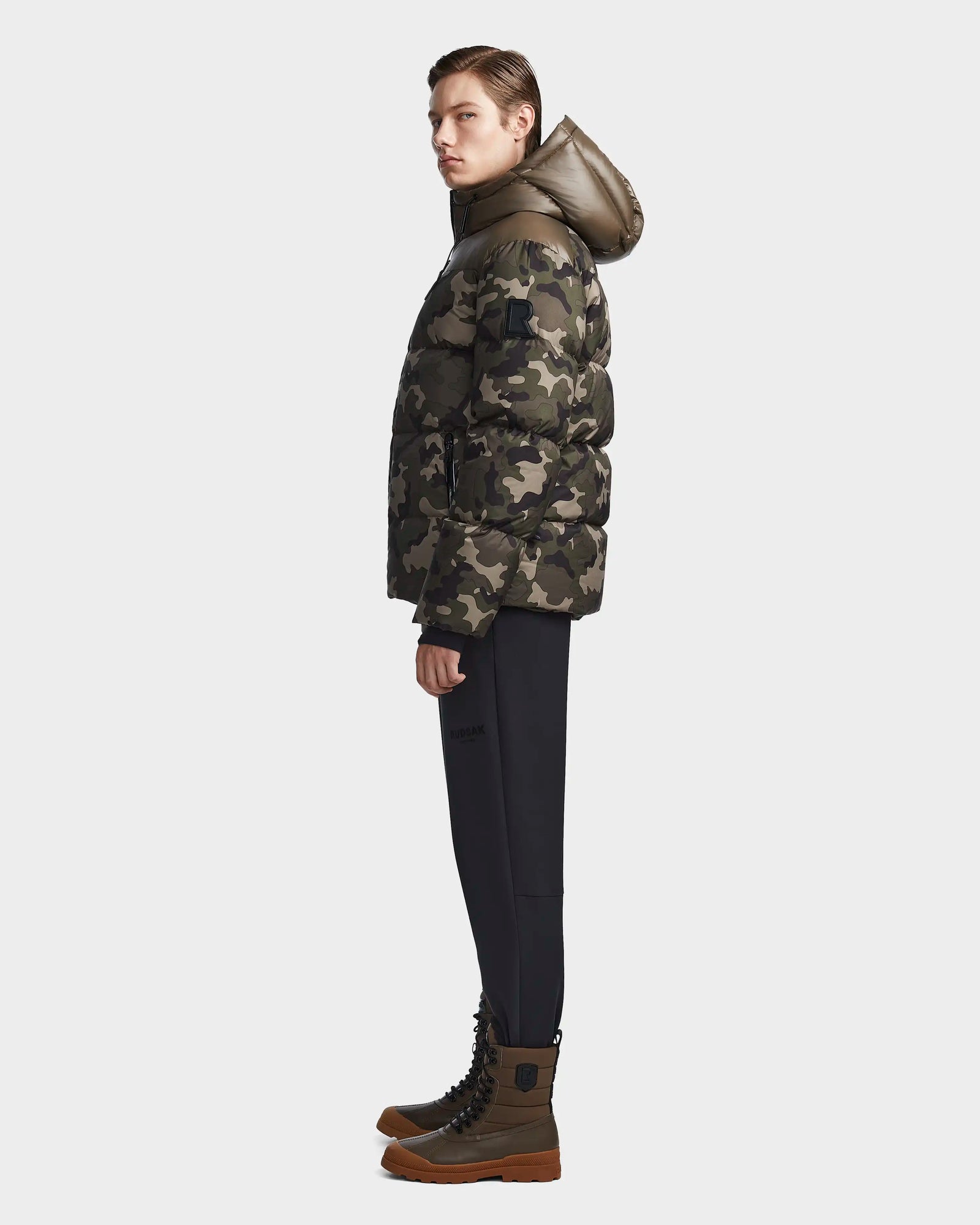 A person stands in profile wearing the Rudsak RONNY M - M123512 Camouflage weather-resistant jacket with a hood, paired with black pants and brown lace-up boots, set against a plain light background.