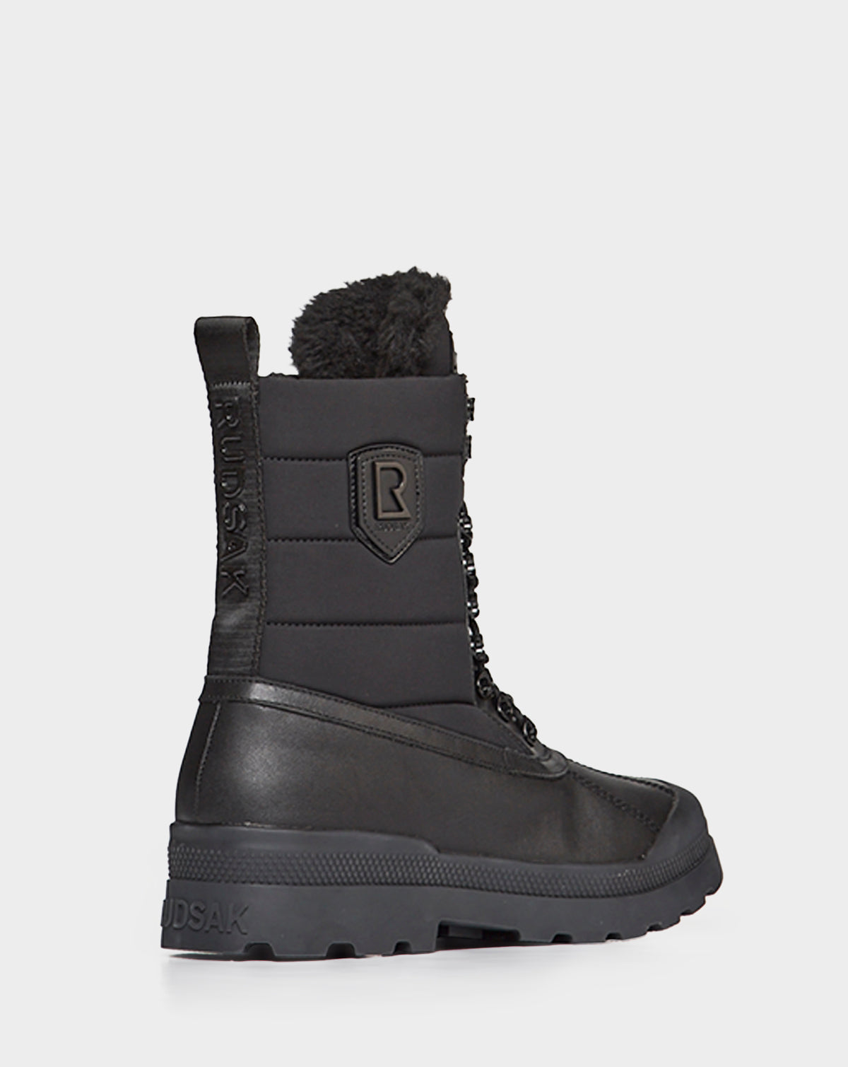 The REAGAN - 6222500 BLACK by Rudsak is a men’s black winter boot with a thick rubber sole, lace-up front, padded upper, faux fur collar, and Rudsak branding on the side and heel—crafted for comfort like premium leather boots.
