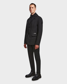 A man stands in front of a plain white background, wearing the Rudsak OTTO - 6124602 Black men’s blazer-style down jacket with eco polyfill, paired with black pants, a turtleneck, and boots.