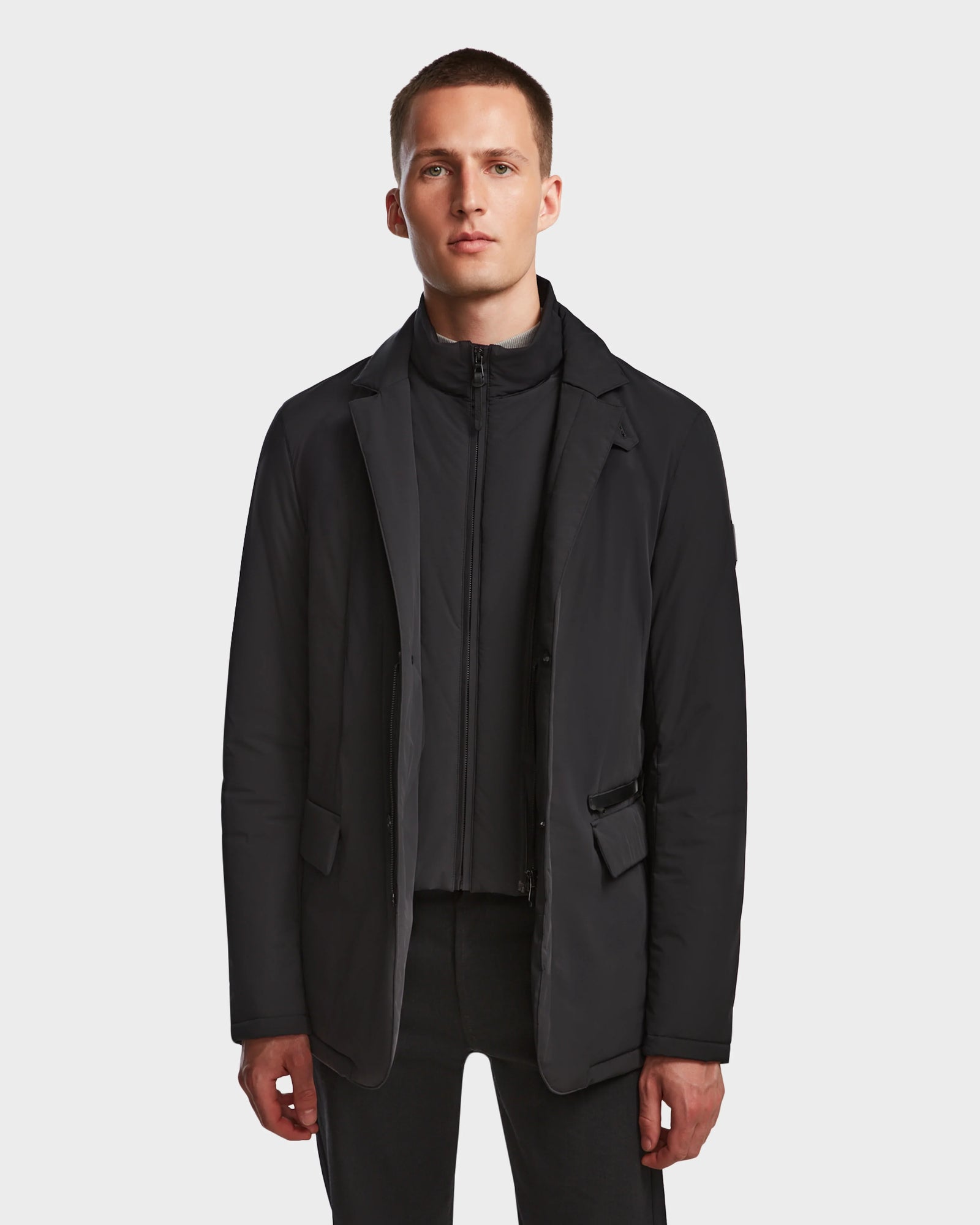 A man with short hair wears the Rudsak OTTO - 6124602 Black vegan leather mens blazer-style down jacket over a black zip-up top and black pants, standing against a plain white background.