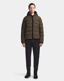 A person stands facing forward, wearing the Rudsak DEMIAN M - M123507 Light Olive down puffer, paired with black pants and lace-up boots against a plain white background.