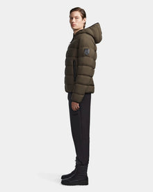 A person stands in profile wearing the Rudsak DEMIAN M - M123507 Light Olive hooded down puffer made from sustainable recycled fabric, paired with black pants and lace-up boots against a plain light background.