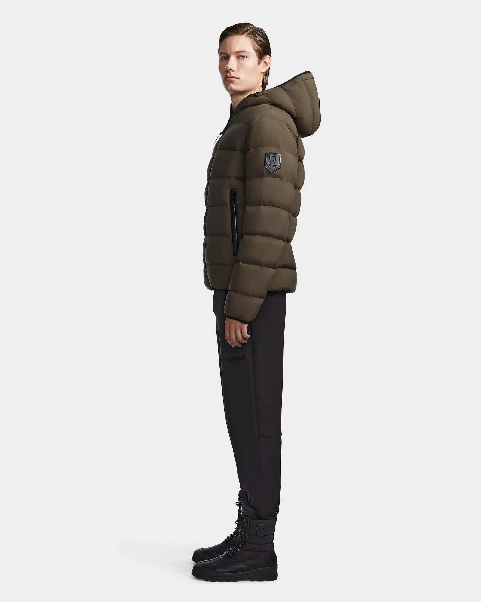 A person stands in profile wearing the Rudsak DEMIAN M - M123507 Light Olive hooded down puffer made from sustainable recycled fabric, paired with black pants and lace-up boots against a plain light background.