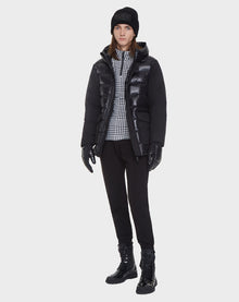 Wearing the Rudsak DUSK - M121506 BLACK men's puffer jacket with waterproof zippers, a black beanie, gloves, pants, and boots. A plaid shirt peeks out underneath. The background is plain white.