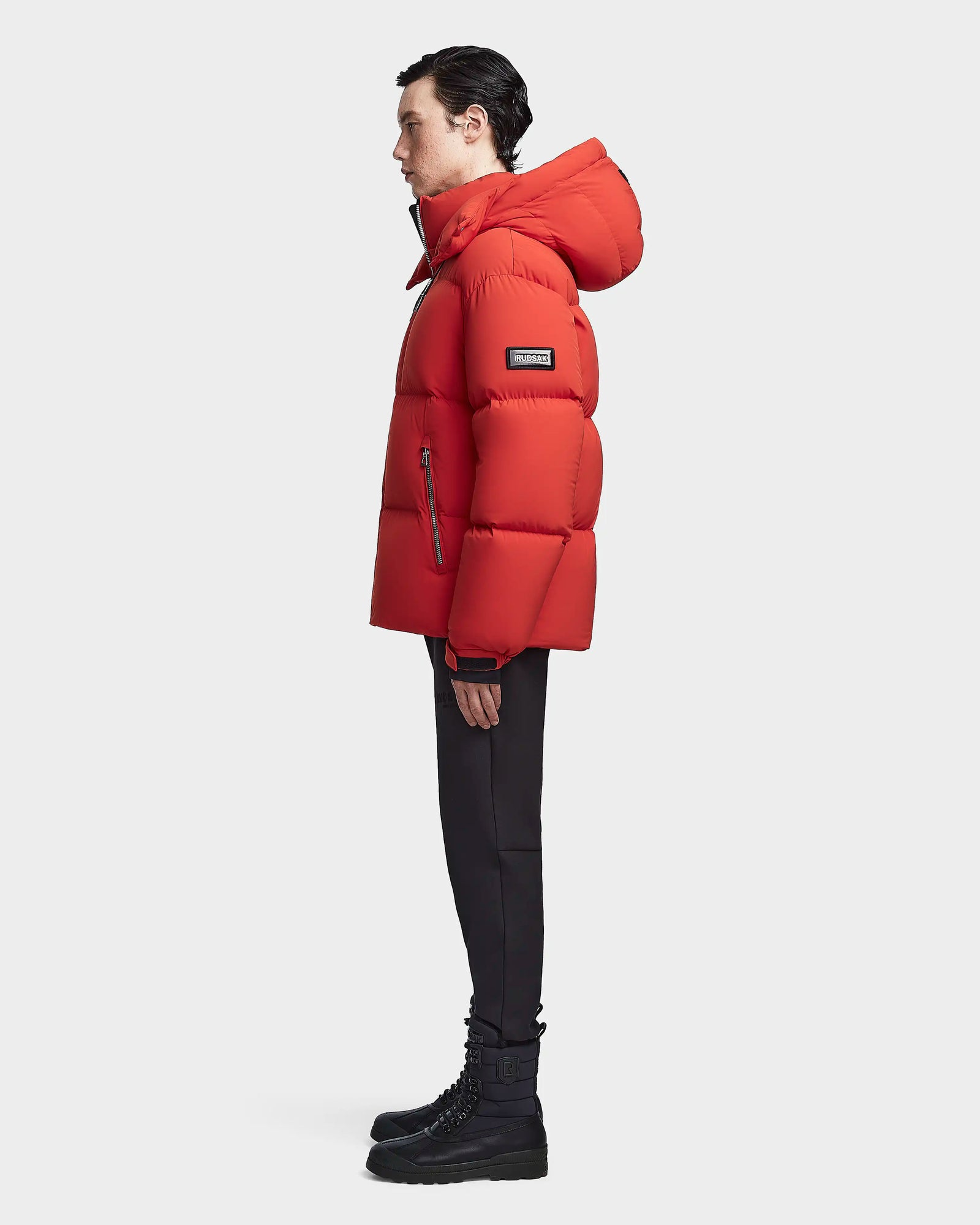 A man in profile wears the Rudsak VINCENT M - 6123510 Orange down puffer jacket, paired with black pants and lace-up boots, standing against a plain white background.