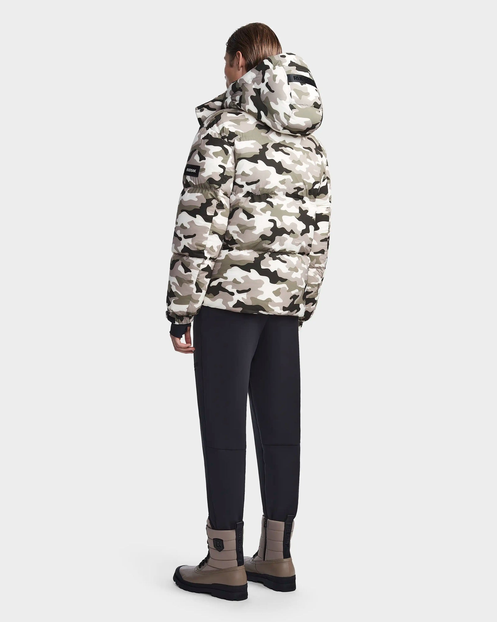 A person stands facing away, wearing the Rudsak VINCENT M - 6123510 Camouflage men's down puffer in white, gray, and black camo with a hood, black pants, and tan boots against a plain light gray background.