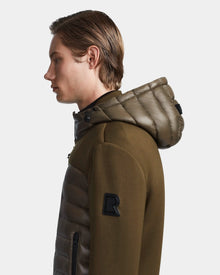A young man with straight, light brown hair is shown in profile wearing the Rudsak DURIN S - M123518 Light Olive men’s hooded jacket with quilted panels, lightweight down insulation, and an “R” patch on the sleeve against a plain background.