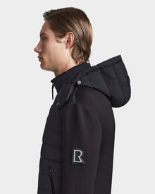 A young man with straight light brown hair is shown in profile wearing the Rudsak LAURENZO - M123514 Black neoprene jacket with quilted pattern, removable hood, and large R patch on the sleeve, against a plain white background.