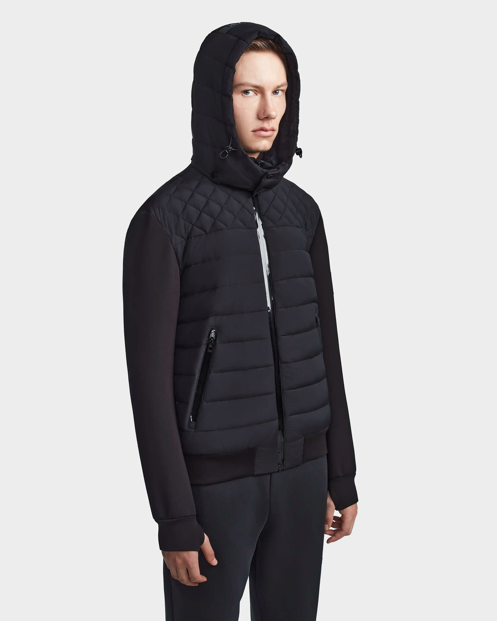 A person wearing the Rudsak LAURENZO - M123514 Black men's light down jacket with removable hood and zippered pockets, paired with black pants, stands slightly to the side against a plain white background.