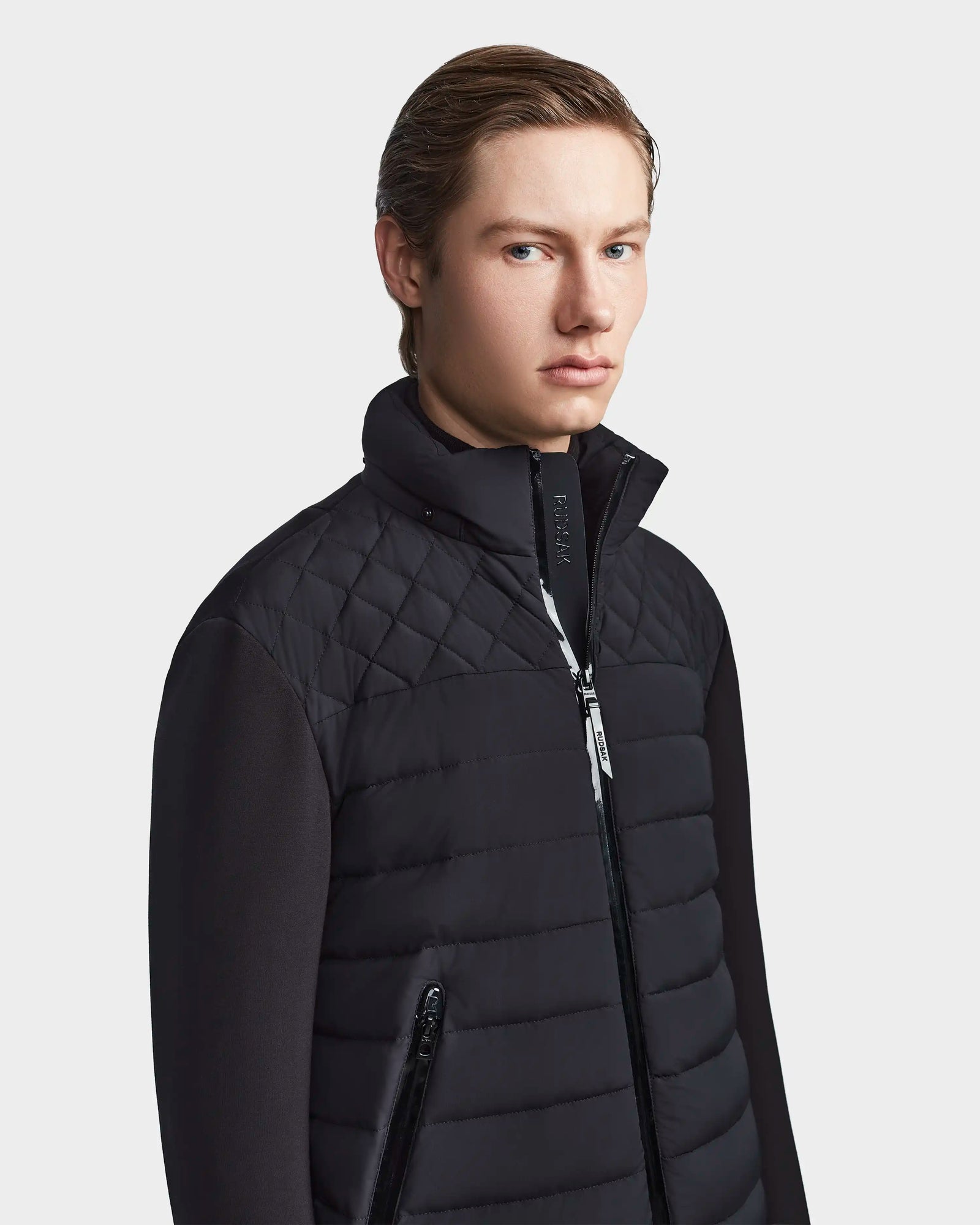 A young person with light skin and slicked-back hair models the Rudsak LAURENZO - M123514 Black men's down jacket with a high collar, looking slightly to the side against a plain light background.