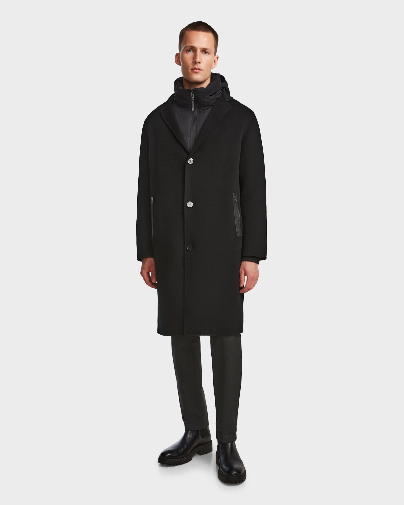 A man faces forward against a white background, wearing Rudsaks DRAL - 6124503 Black 2-in-1 jacket with a removable hooded down puffer, dark pants, and black boots.