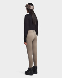 A woman with long dark hair stands facing away, wearing Rudsaks ARIL M - 8522514 Cement sustainable winter pants in beige, a black sweater, black platform shoes, and a taupe fur headband against a plain background.